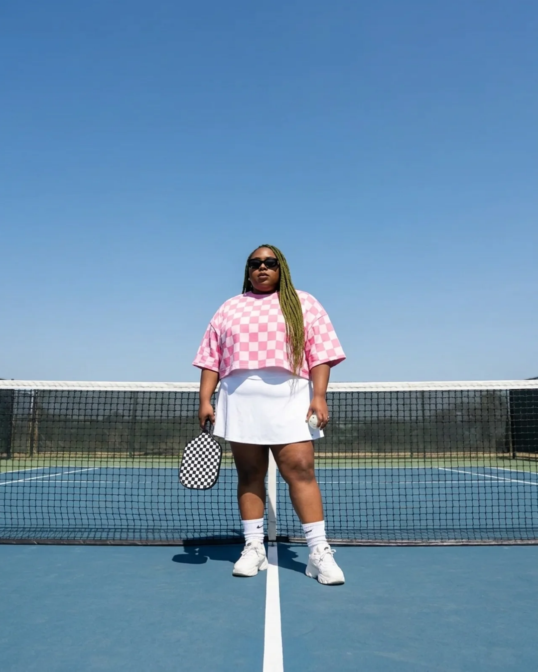 Woman standing on a pickleball court holding a checkered paddle and ball, wearing a pink checkerboard shirt and white skirt